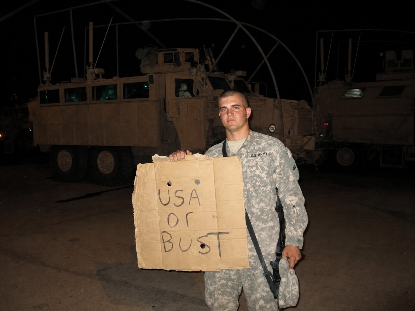 Johnathan Melin, U.S. Army veteran and now Idaho REALTOR® at Good News Realty Group, in ACU uniform holding a 'USA OR BUST' cardboard sign in front of MRAP vehicles at the end of his Iraq deployment, 2009 — Operation Iraqi Freedom