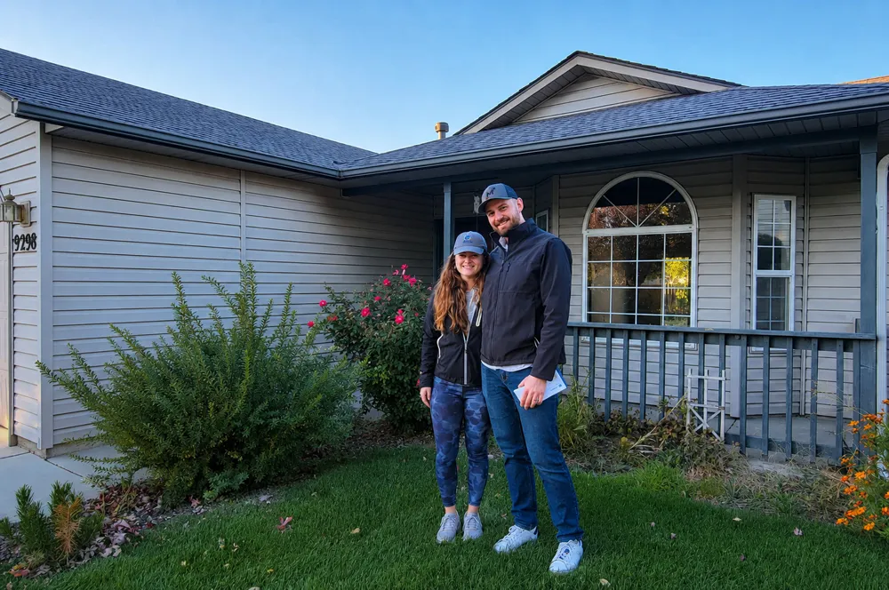 Two new homeowners standing in the front yard of their single-story home at dusk