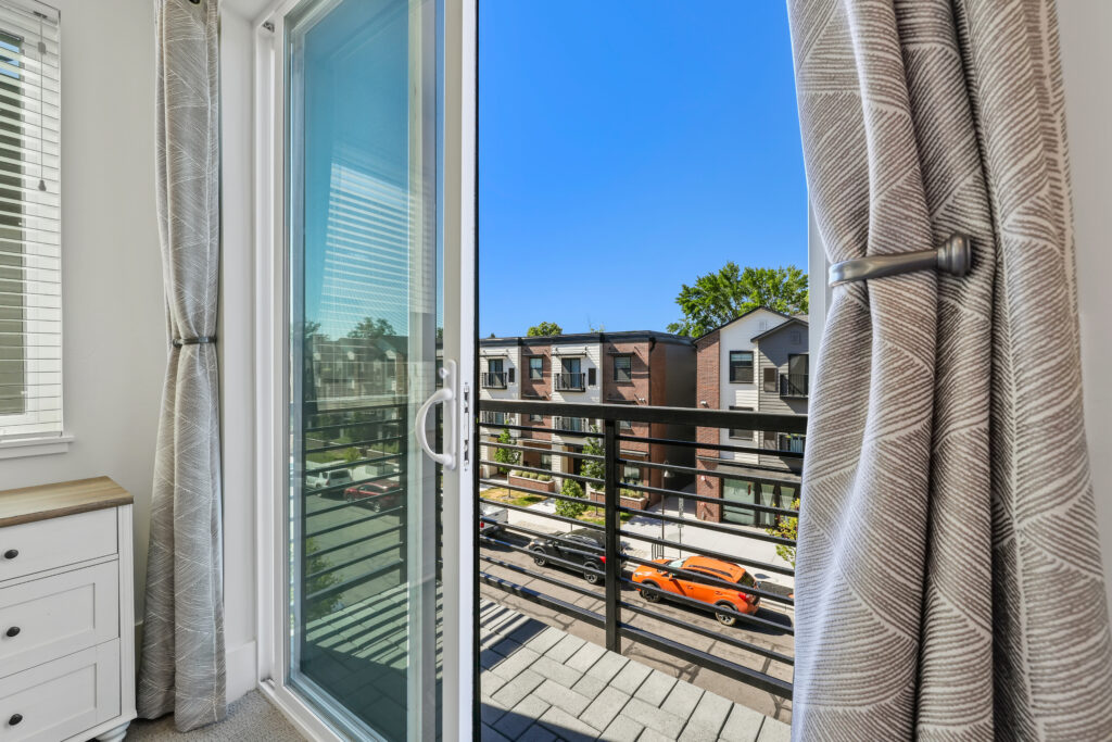 Balcony View from Primary Suite - Good News Realty Group View from bedroom balcony over a street with neighboring townhomes and blue skies.